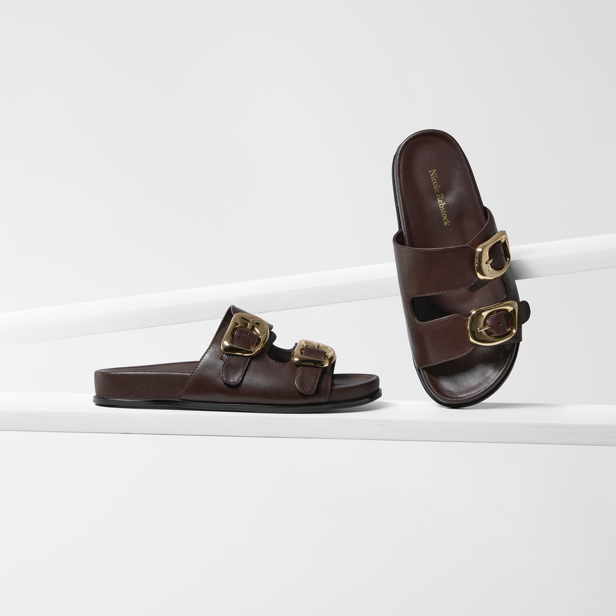 A pair of dark brown leather slide sandals with double gold-tone buckle straps, displayed on a clean white geometric riser.