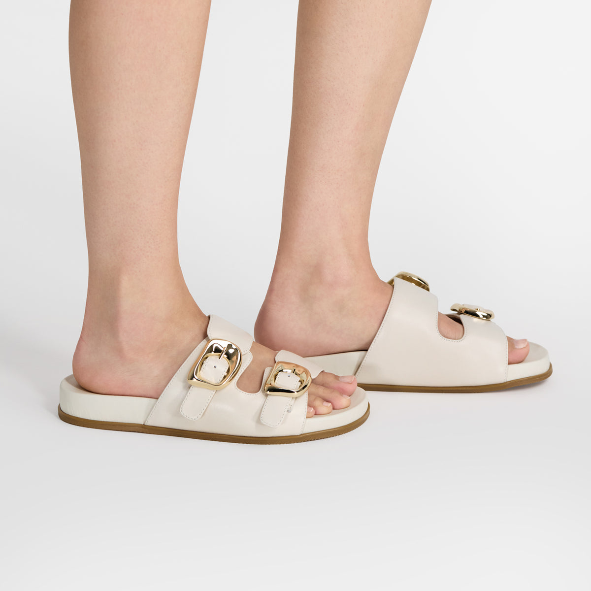 A pair of ivory leather double-strap slide sandals with gold-tone buckle details, shown on a model's feet against a clean white background.