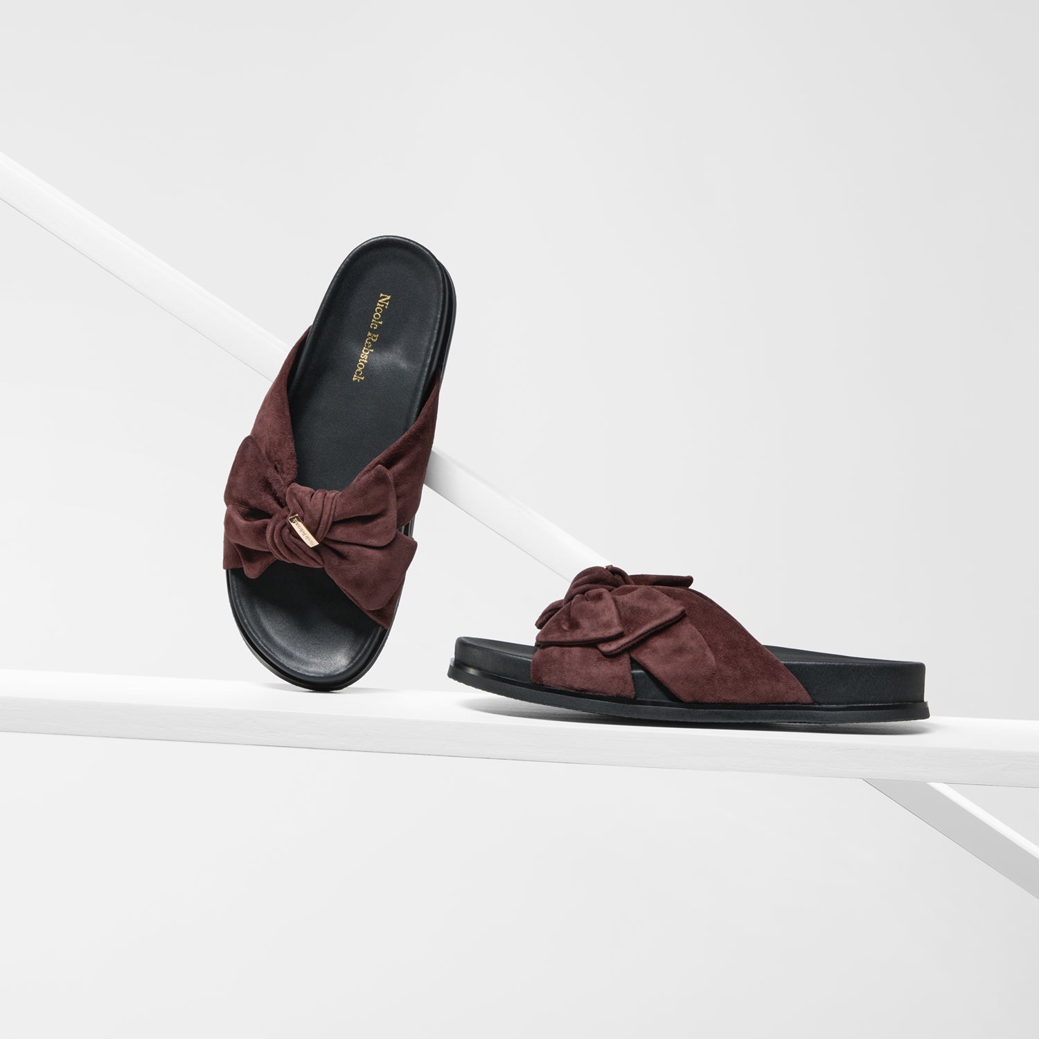 A pair of women's slide sandals in dark brown suede and black, featuring a large bow detail on the strap and a small gold-tone metal accent, displayed on a clean white geometric set.