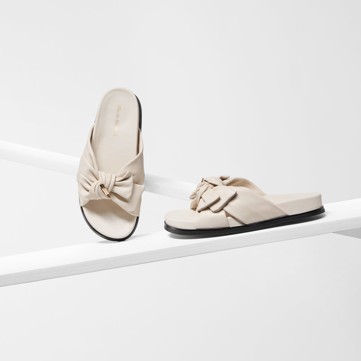 A pair of ivory slide sandals with large bow details on the straps, displayed on a clean white geometric riser against a white background.