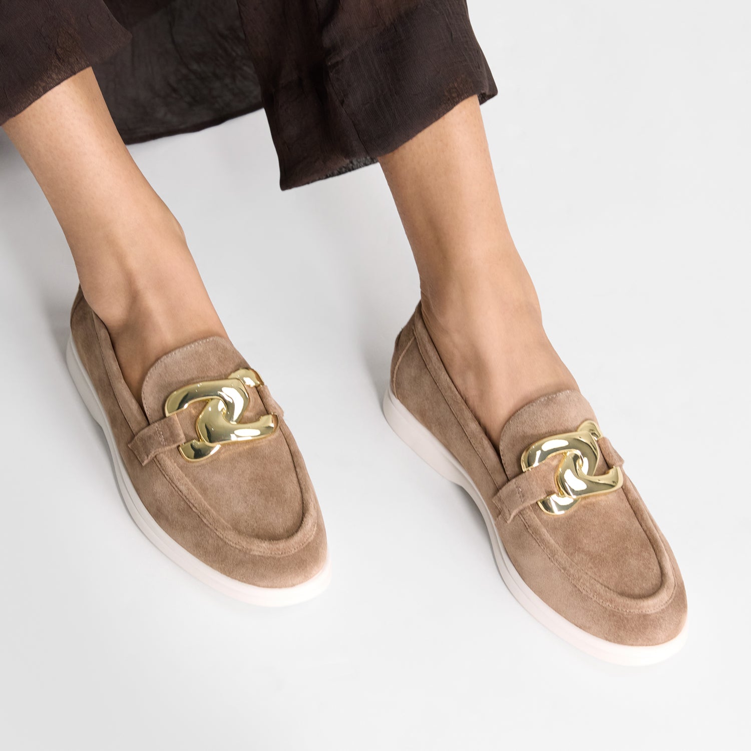Beige  suede loafers with gold accents worn by a person on a light grey background
