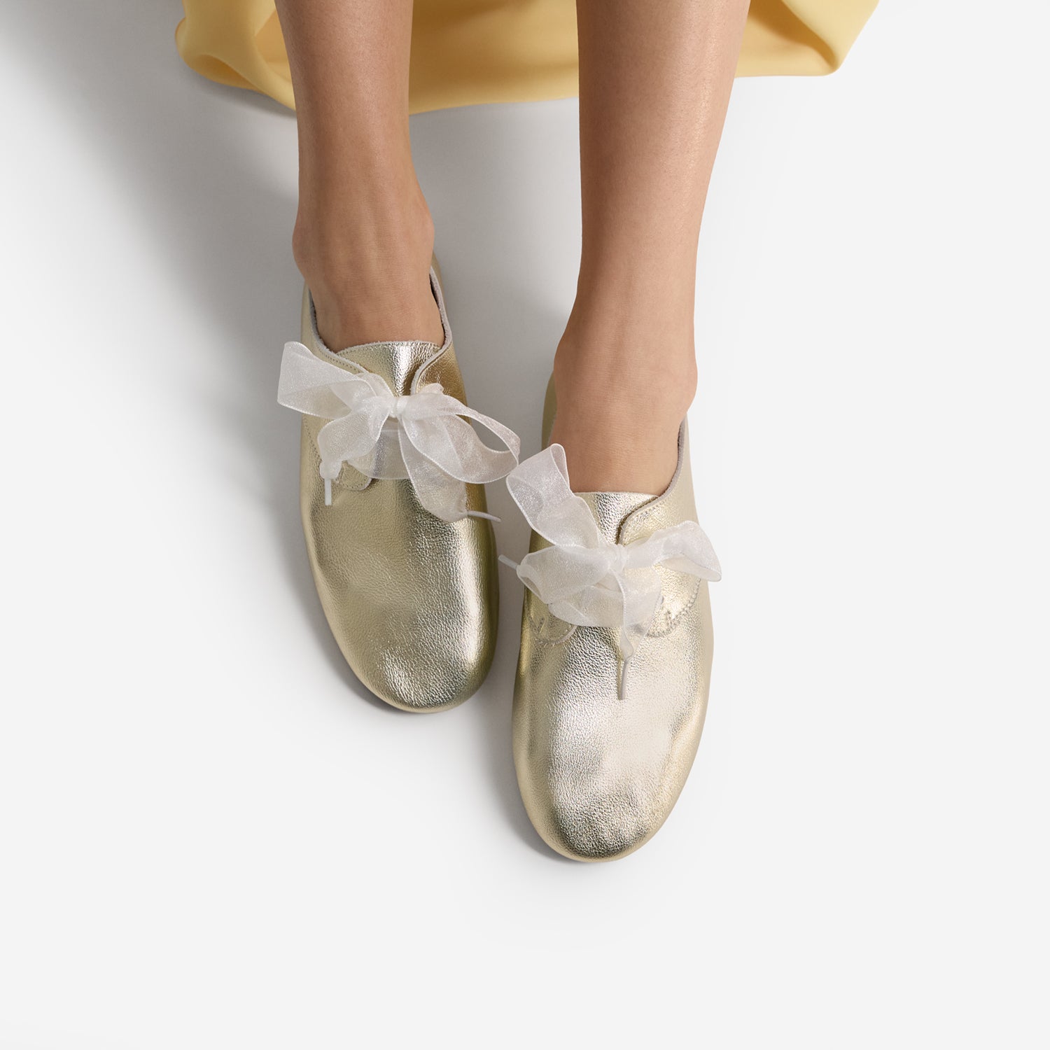Gold shoes called Xeno with white ribbons worn by a person on a light gray background
